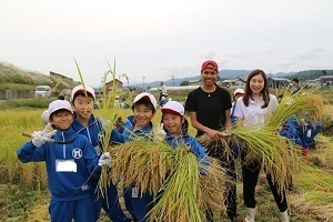 国際大学の学生と児童が刈り取った稲をもって笑顔の様子
